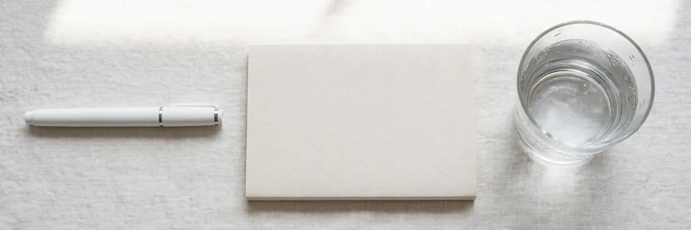 White pen, blank notebook and a glass of water on a light cloth, seen from above